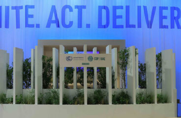 Detail of the COP28’s Trade House pavilion in Dubai, United Arab Emirates. Background: a soft blue curtain with the words “UNITE. ACT. DELIVER”. Foreground: a stage with plants and a big white poster with the printed logos of the UNFCC and the COP28 UAE