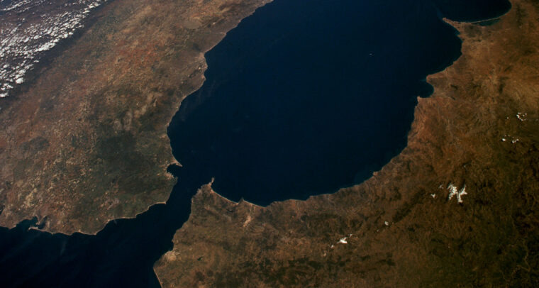 Daytime aerial view of the Strait of Gibraltar, separating Spain (north) and Morocco (south). Visible features include the Sierra Nevada in Spain, the mountainous coastline of Morocco, the River Guadalquivir, and the cities of Gibraltar and Ceuta under clear skies. Ceuta and Melilla