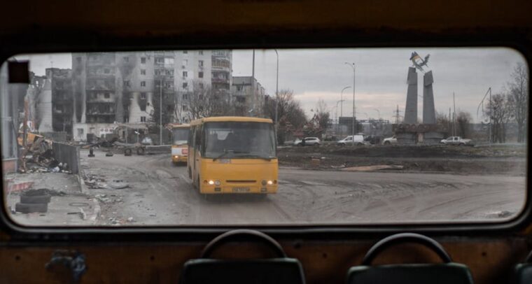 A view from inside an old yellow bus shows a war-torn cityscape in Borodyanka, Ukraine. Through the rear window, two yellow buses are driving along a muddy road, passing a heavily damaged block of flats. In the background, a monument with a Ukrainian flag on top can be seen. Ukraine