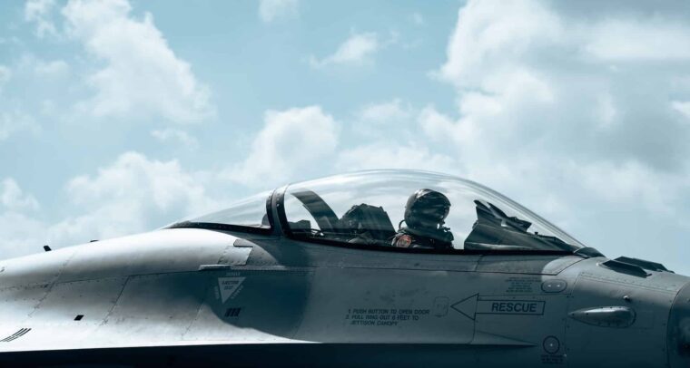 A Belgian F-16 pilot, wearing a flight helmet, oxygen mask, and full flight suit, sits inside the bubble canopy of a grey F-16 Fighting Falcon. The cockpit features visible instrument panels and side-stick controls, with warning labels on the fuselage just below the cockpit. The aircraft is parked on the tarmac at Florennes Air Base under a bright, partly cloudy sky during a NATO Quick Reaction Alert (QRA) exercise. Defence