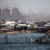 A line of tents near the port of Gaza, with several people gathered along the shoreline. In the background, buildings can be seen with smoke rising behind them. In the foreground, a partially submerged structure is visible in the water. Gaza