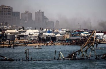 A line of tents near the port of Gaza, with several people gathered along the shoreline. In the background, buildings can be seen with smoke rising behind them. In the foreground, a partially submerged structure is visible in the water. Gaza